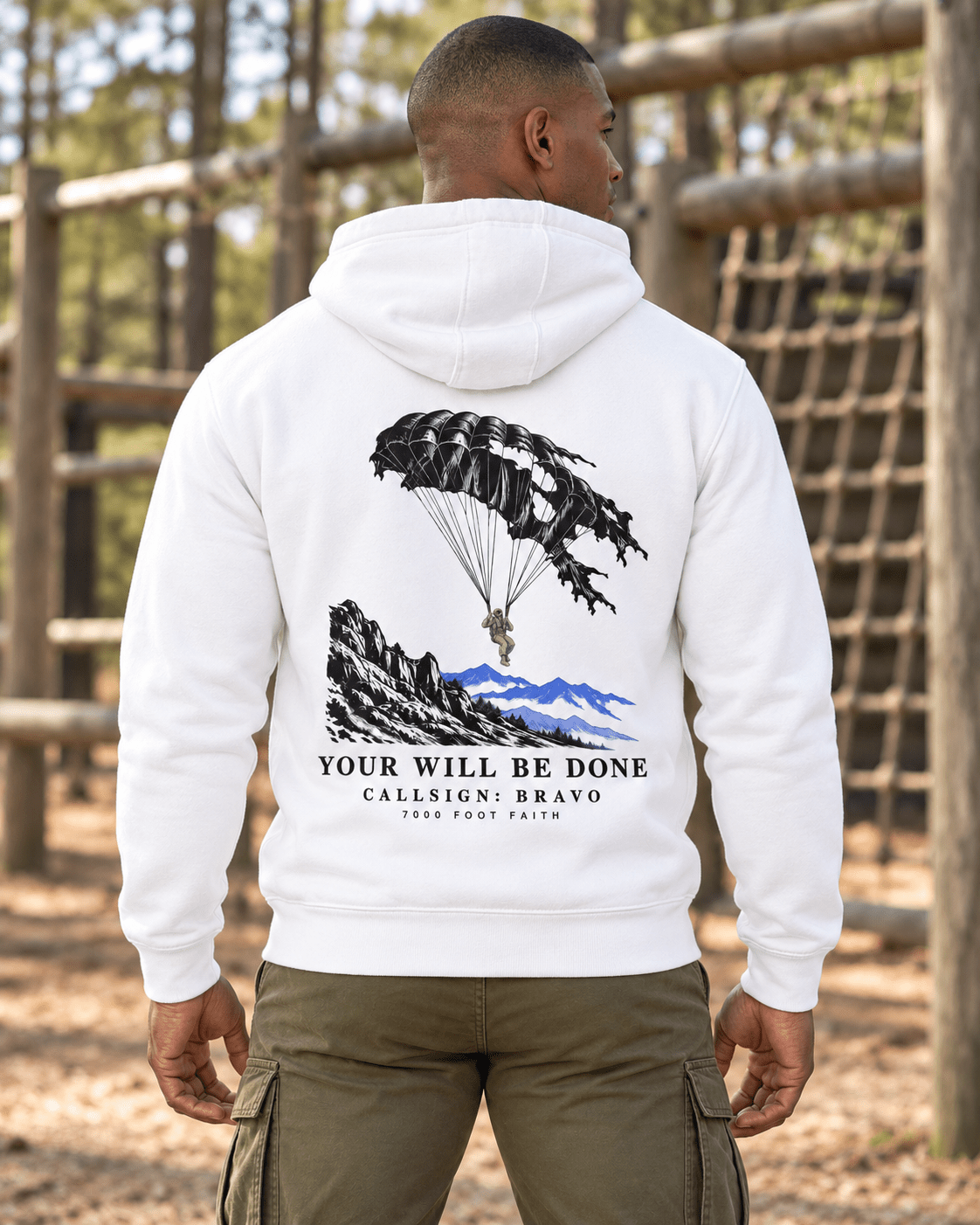 Man at an outdoor training course wearing a White DESCENT faith hoodie, back view showing parachute and mountain artwork with Your Will Be Done lettering