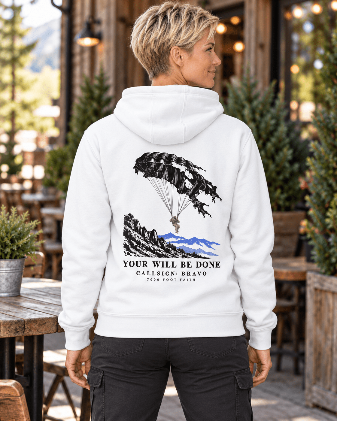 Woman on a cafe patio wearing a White DESCENT faith hoodie, back view showing parachute and mountain artwork with Your Will Be Done lettering