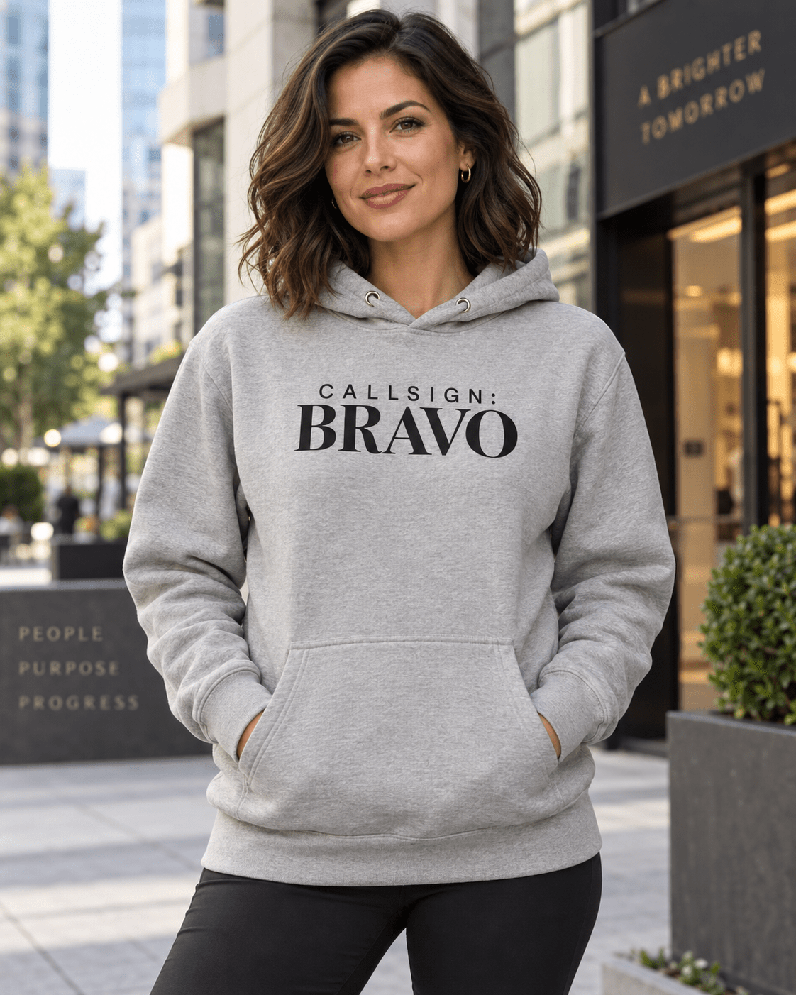 Woman wearing a Carbon Grey God Is Good CALLSIGN: BRAVO hoodie outside a city storefront — front view