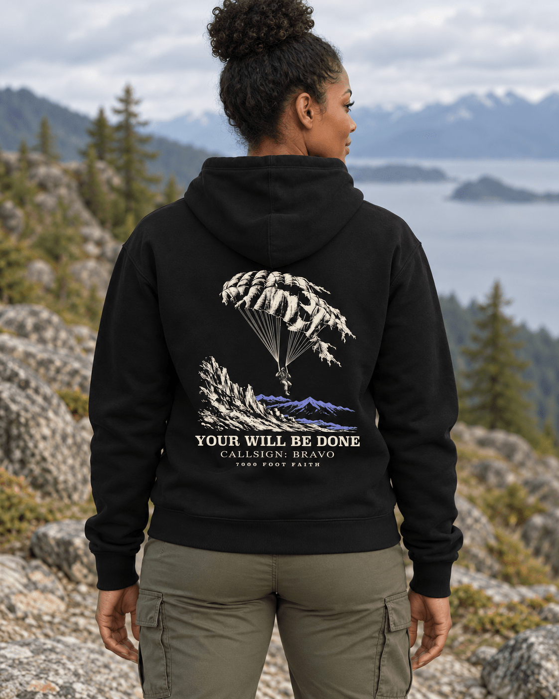 Woman outdoors wearing a Black DESCENT faith hoodie, back view showing parachute and mountain artwork with Your Will Be Done lettering