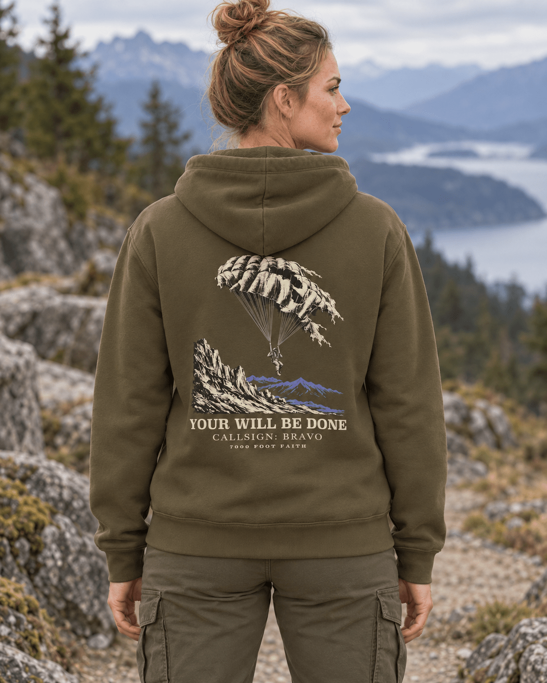 Woman outdoors wearing a Military Green DESCENT faith hoodie, back view showing parachute and mountain artwork with Your Will Be Done lettering