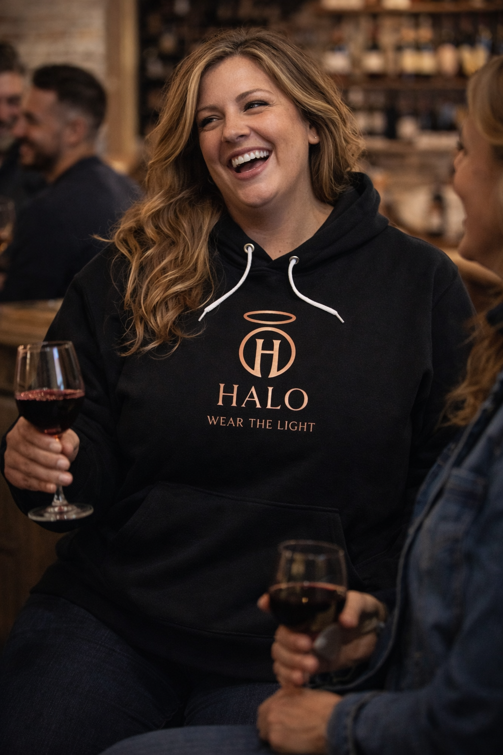Person wearing a black hoodie with 'HALO' logo, holding a glass of red wine in a social setting.