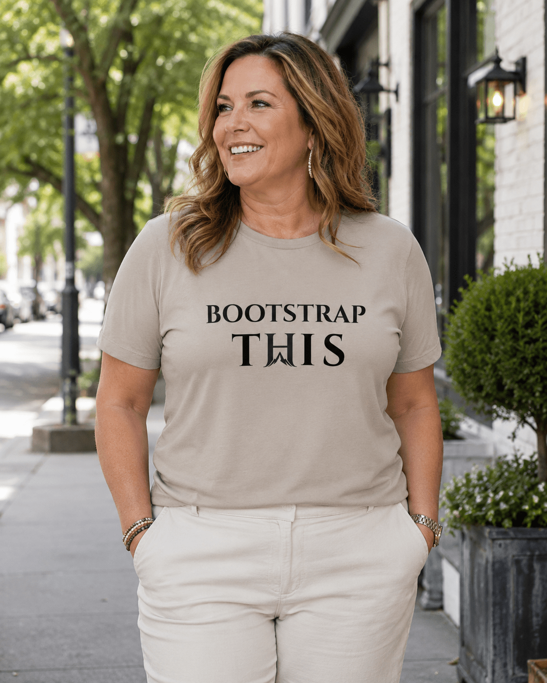 Woman wearing the Heather Stone Bootstrap This Women’s Relaxed Tee with black artwork on a tree-lined sidewalk