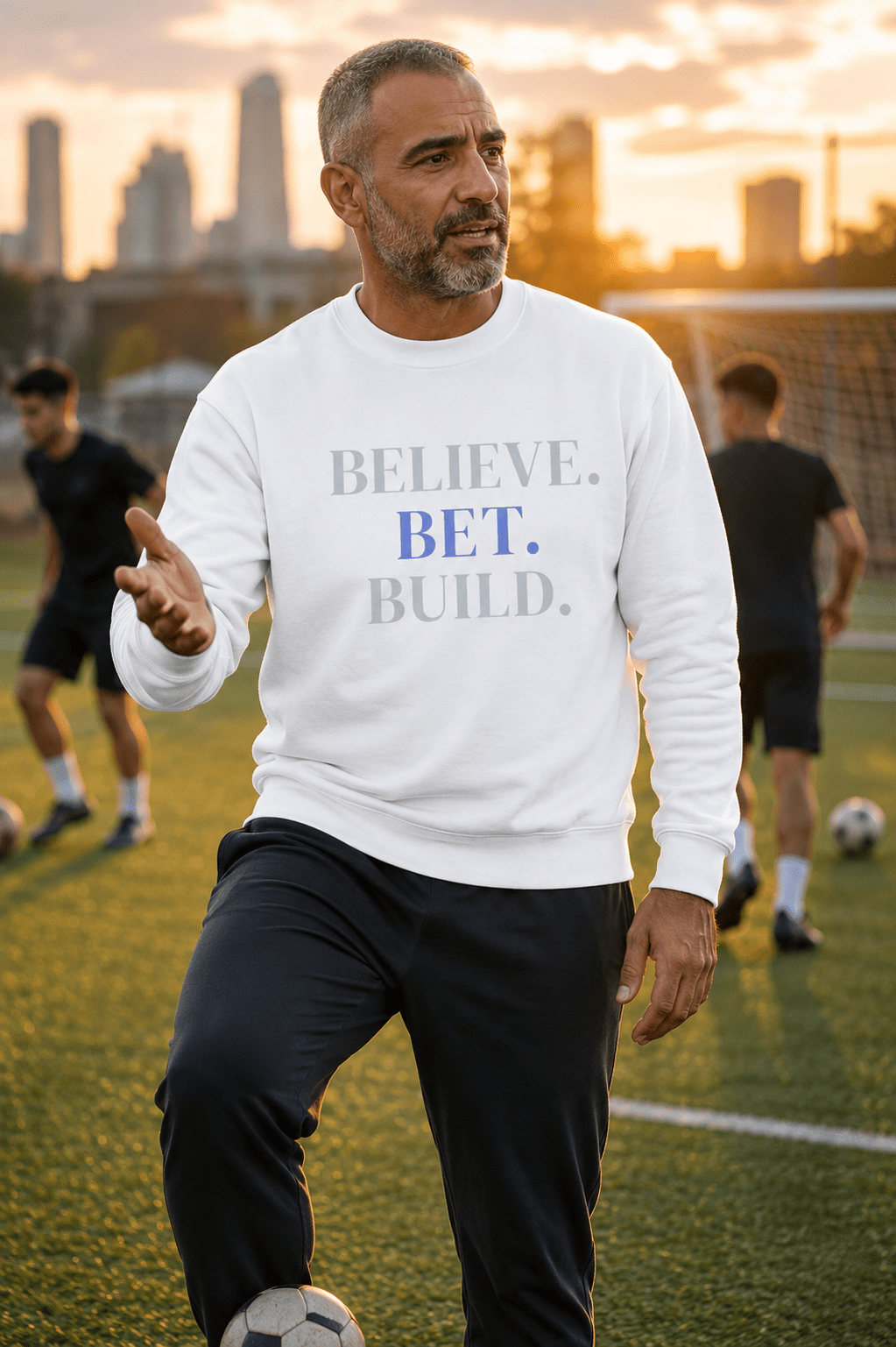 White Bootstrappin’ BELIEVE. BET. BUILD. crewneck — model on a sports field