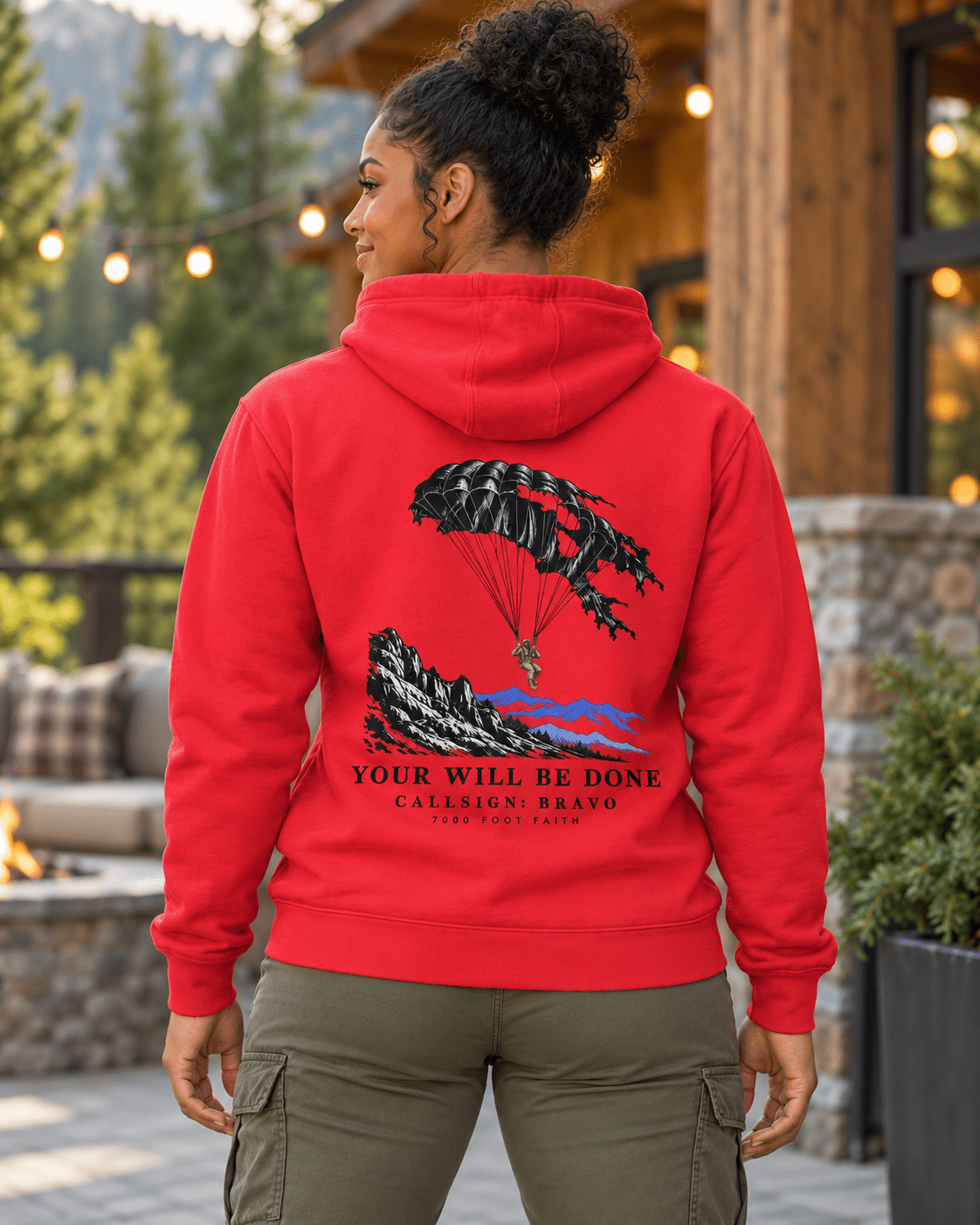 Woman on a lodge patio wearing a Team Red DESCENT faith hoodie, back view showing parachute and mountain artwork with Your Will Be Done lettering