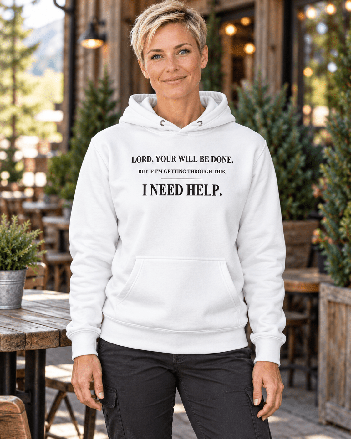 Woman on a cafe patio wearing a White DESCENT faith hoodie, front view showing Your Will Be Done and I Need Help lettering