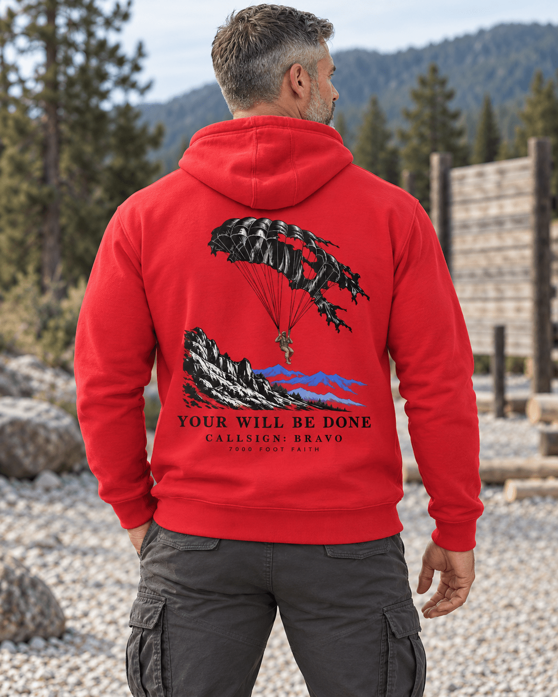 Man at a rugged trailhead wearing a Team Red DESCENT faith hoodie, back view showing parachute and mountain artwork with Your Will Be Done lettering