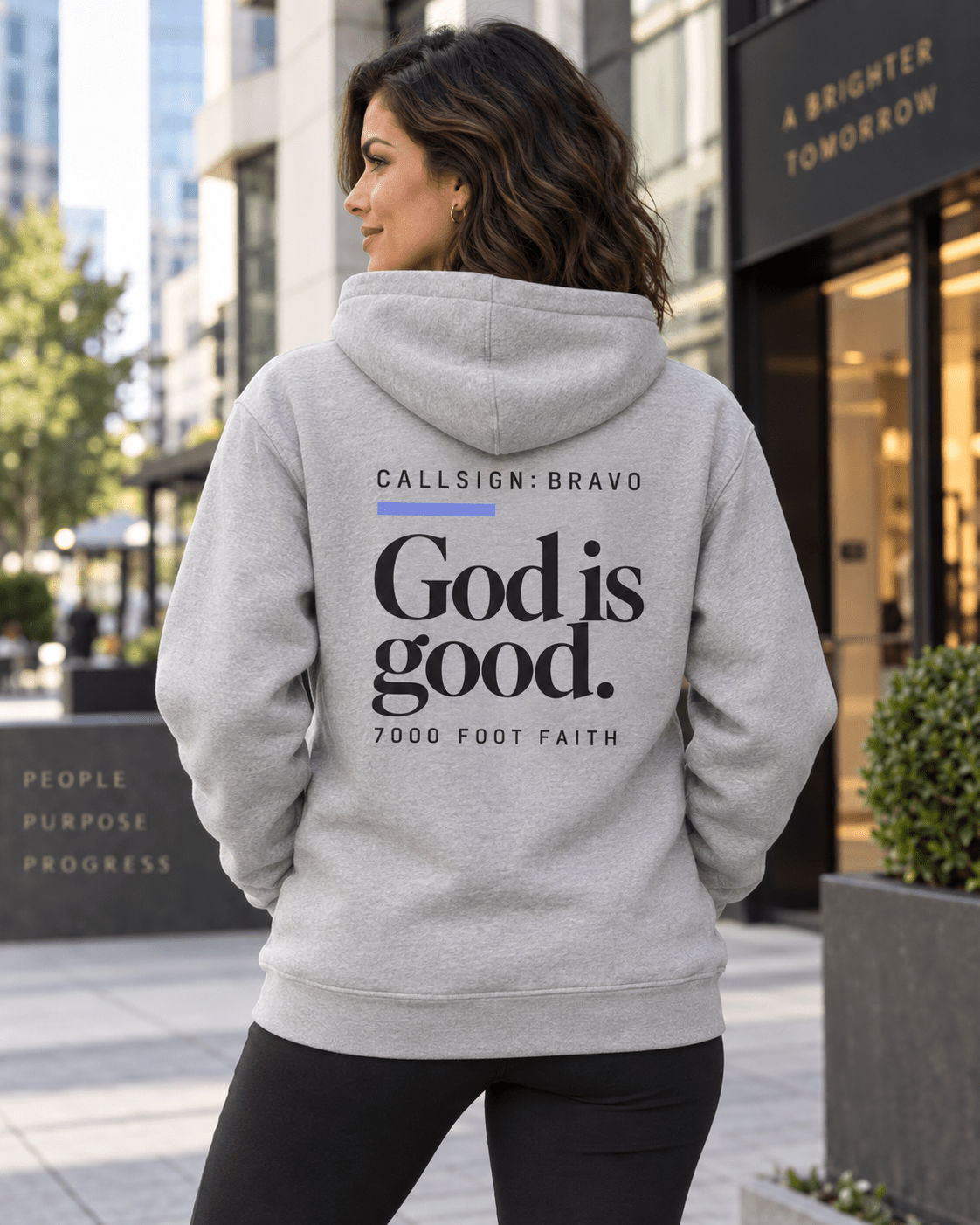 Woman wearing a Carbon Grey God Is Good CALLSIGN: BRAVO hoodie outside a city storefront — back view