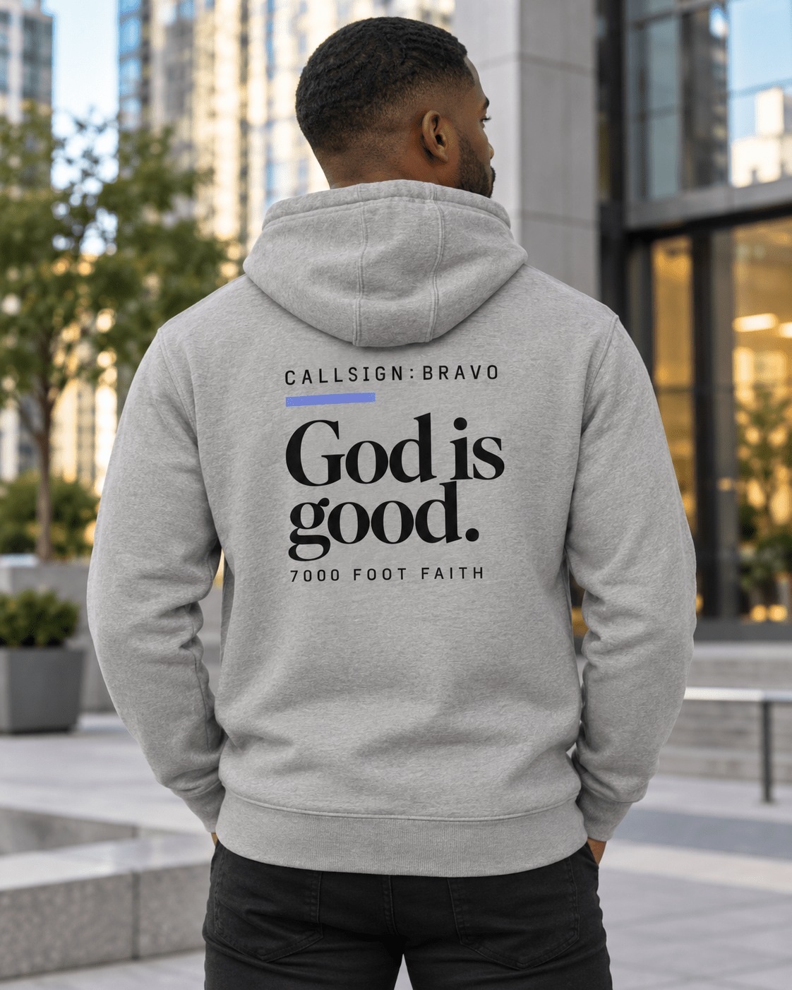 Man wearing a Carbon Grey God Is Good CALLSIGN: BRAVO hoodie in an urban plaza — back view