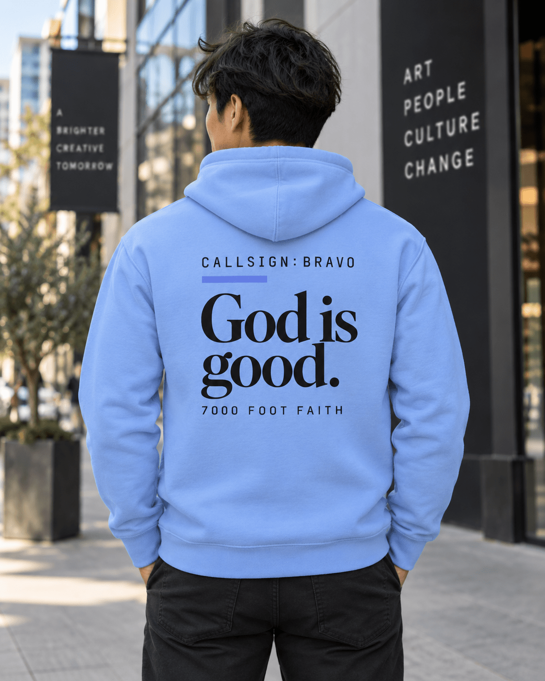Man wearing a Carolina Blue God Is Good CALLSIGN: BRAVO hoodie outside an art gallery — back view