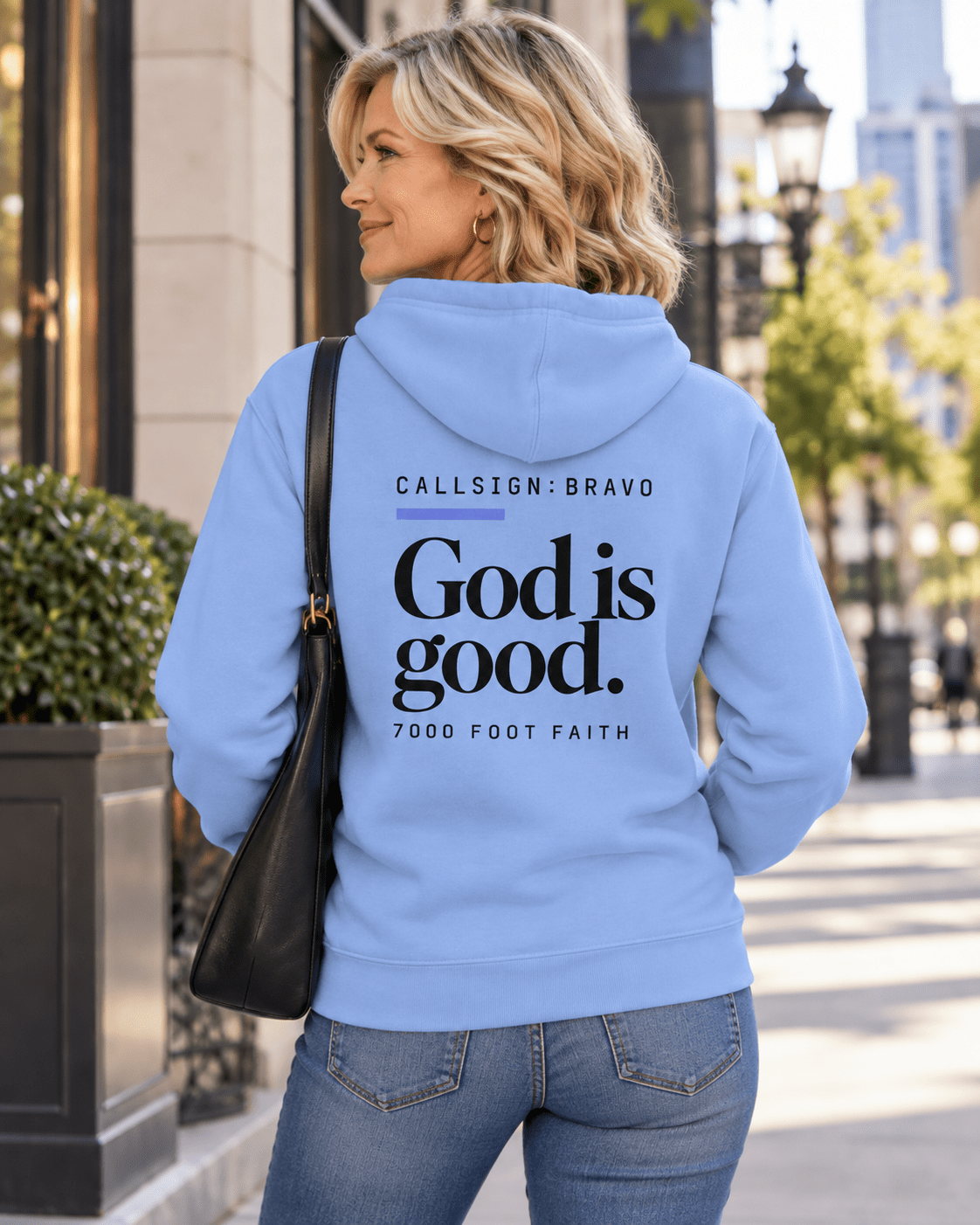 Woman wearing a Carolina Blue God Is Good CALLSIGN: BRAVO hoodie outside a city storefront — back view