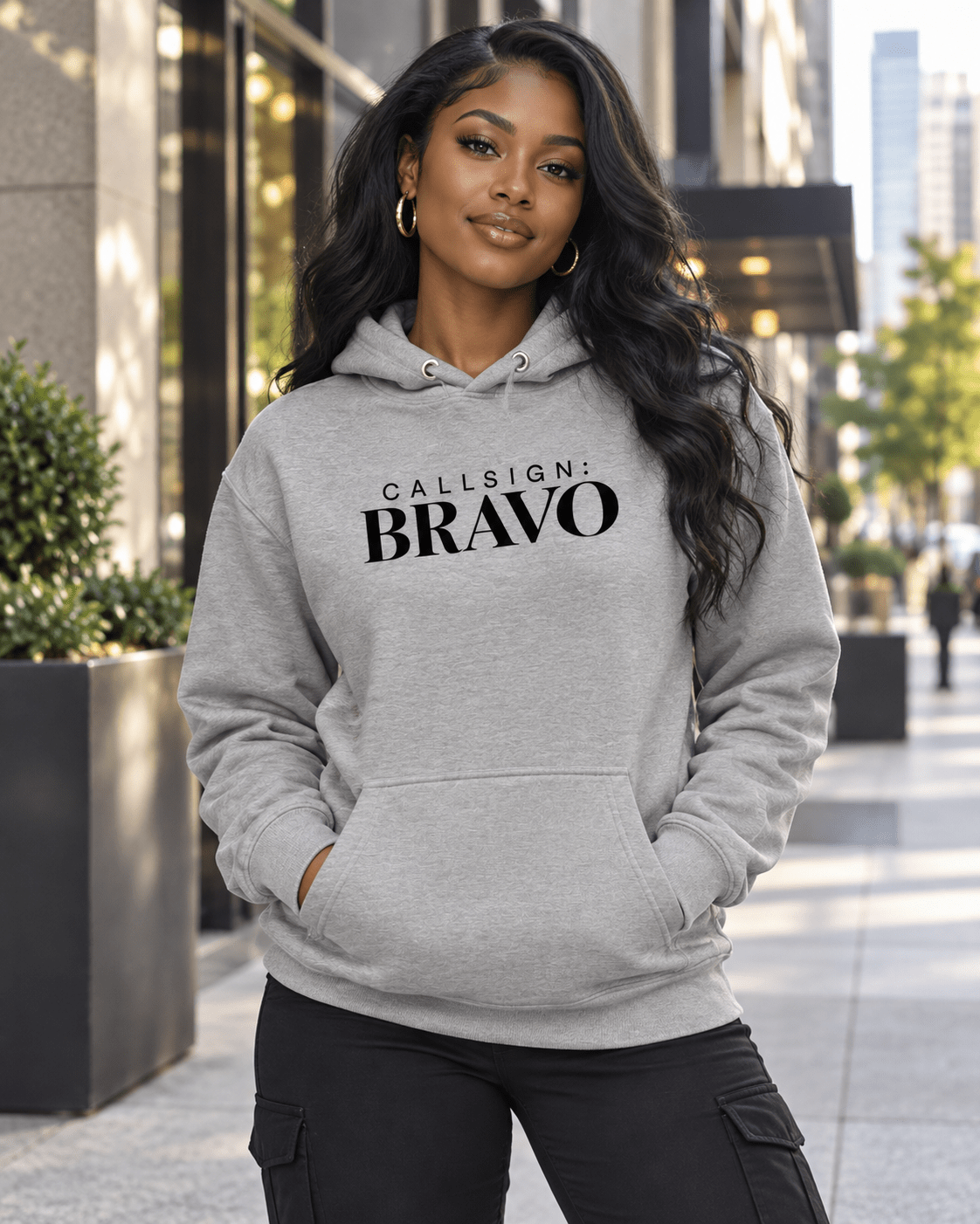 Woman wearing a Carbon Grey God Is Good CALLSIGN: BRAVO hoodie on a city sidewalk — front view