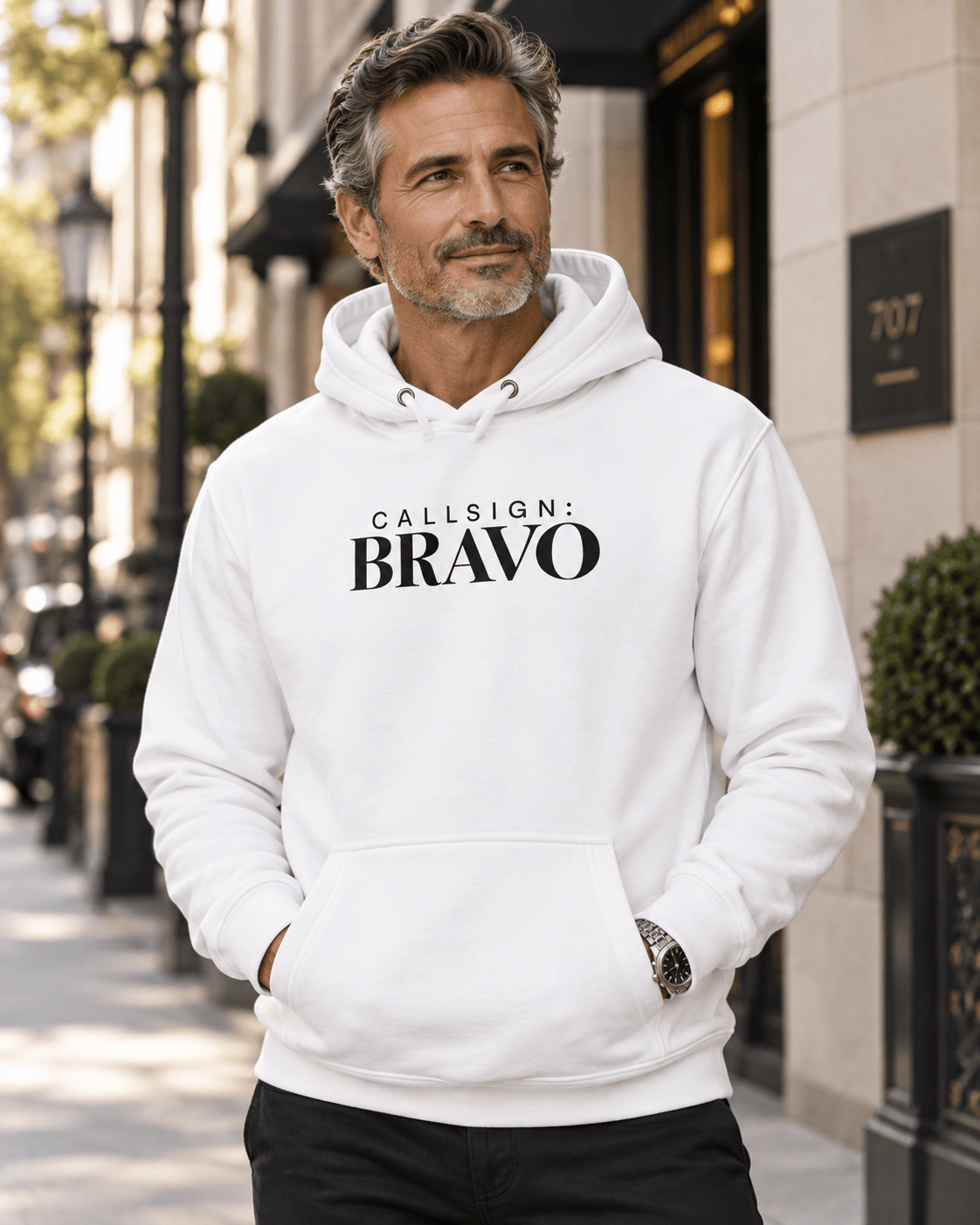 Man wearing a White God Is Good CALLSIGN: BRAVO hoodie beside a city building — front view
