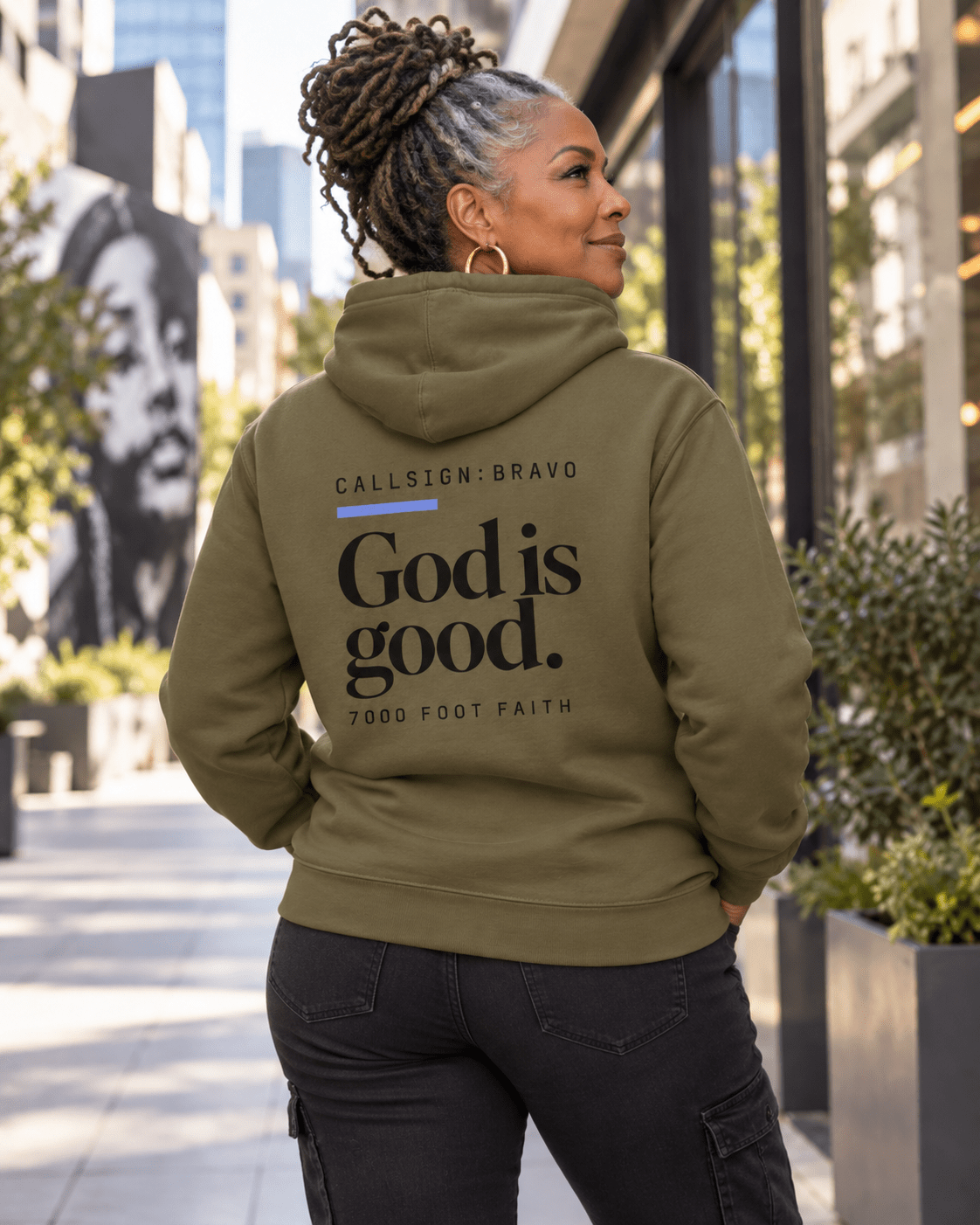 Woman wearing a Military Green God Is Good CALLSIGN: BRAVO hoodie on a city sidewalk — back view