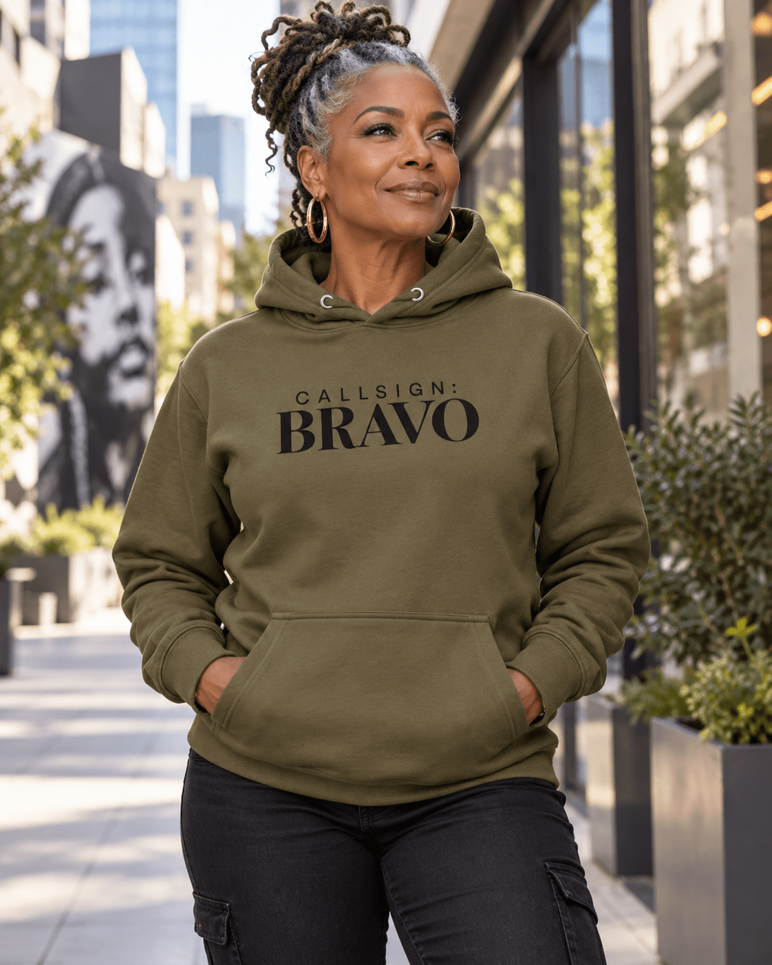 Woman wearing a Military Green God Is Good CALLSIGN: BRAVO hoodie on a city sidewalk — front view