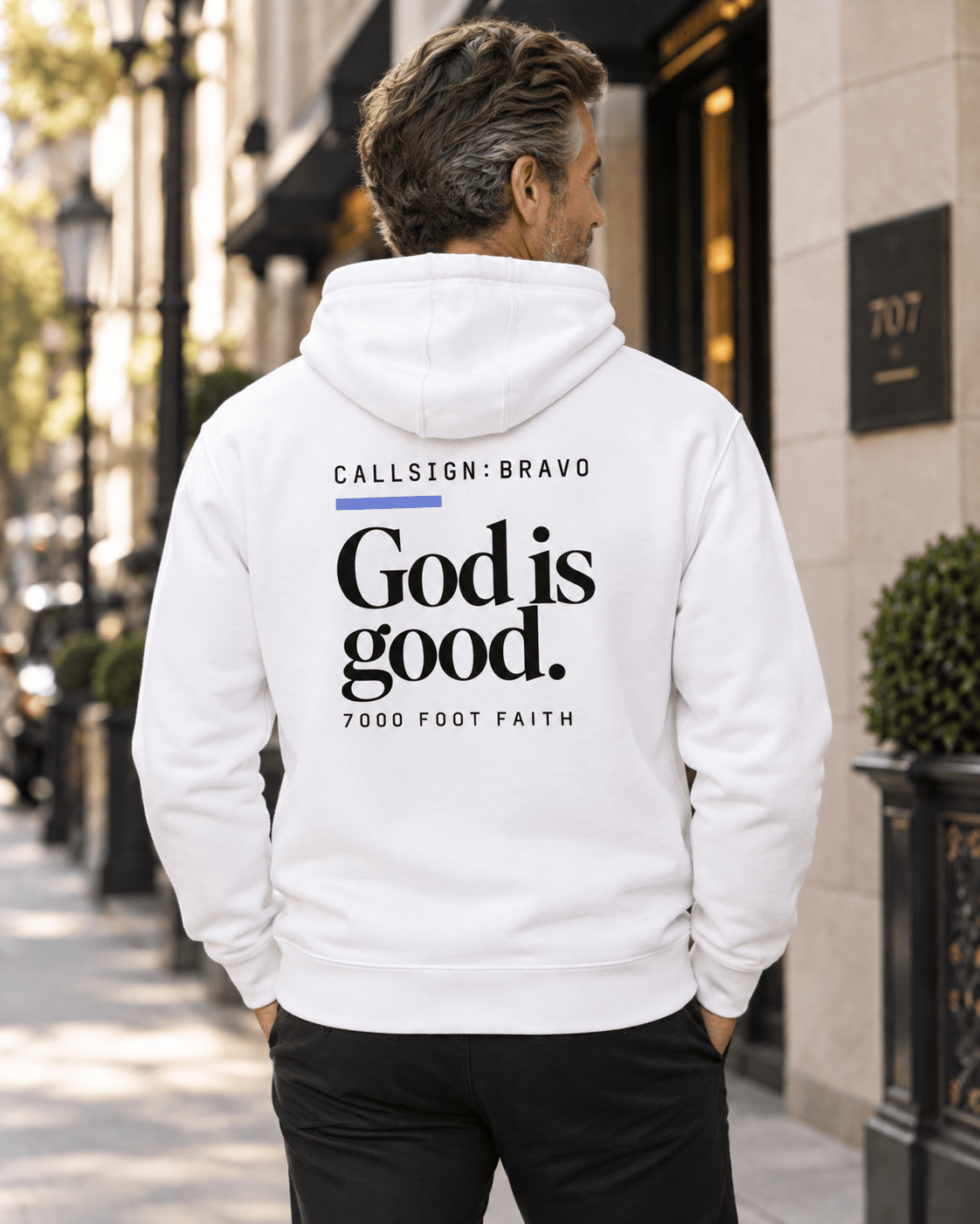 Man wearing a White God Is Good CALLSIGN: BRAVO hoodie beside a city building — back view