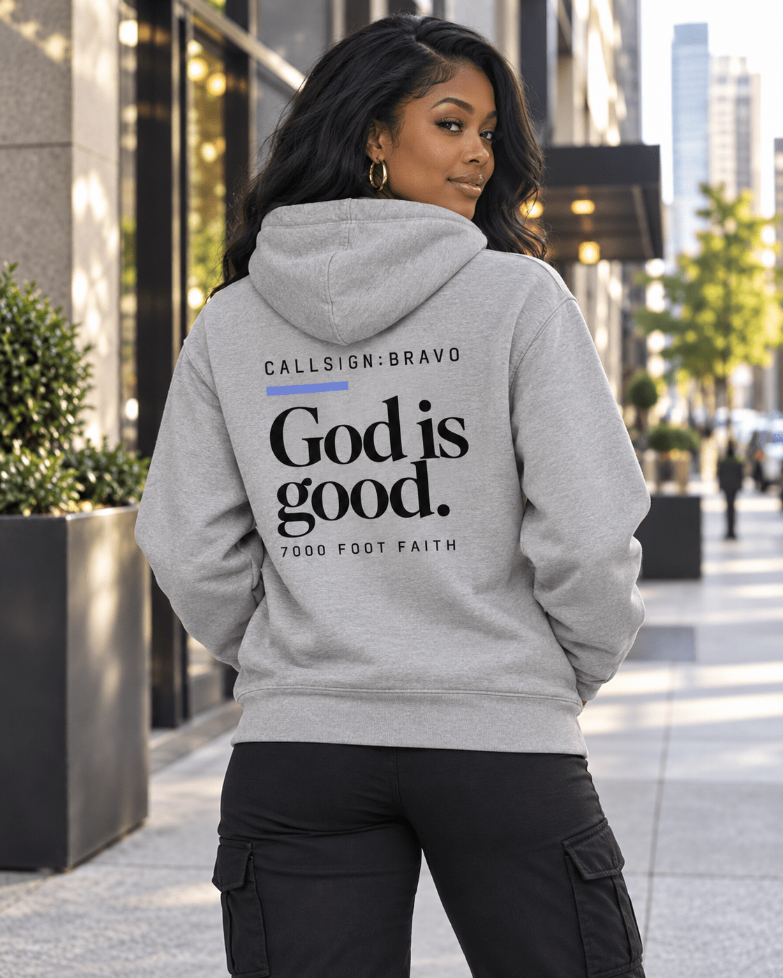 Woman wearing a Carbon Grey God Is Good CALLSIGN: BRAVO hoodie on a city sidewalk — back view
