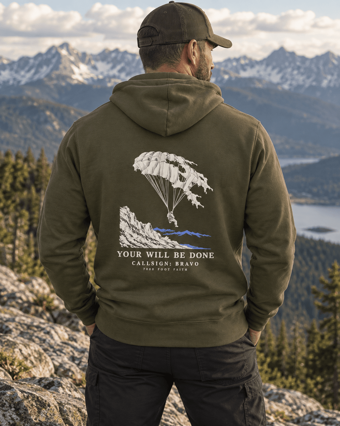 Man wearing a Military Green Your Will Be Done: Descent CALLSIGN: BRAVO hoodie above a mountain lake — back view