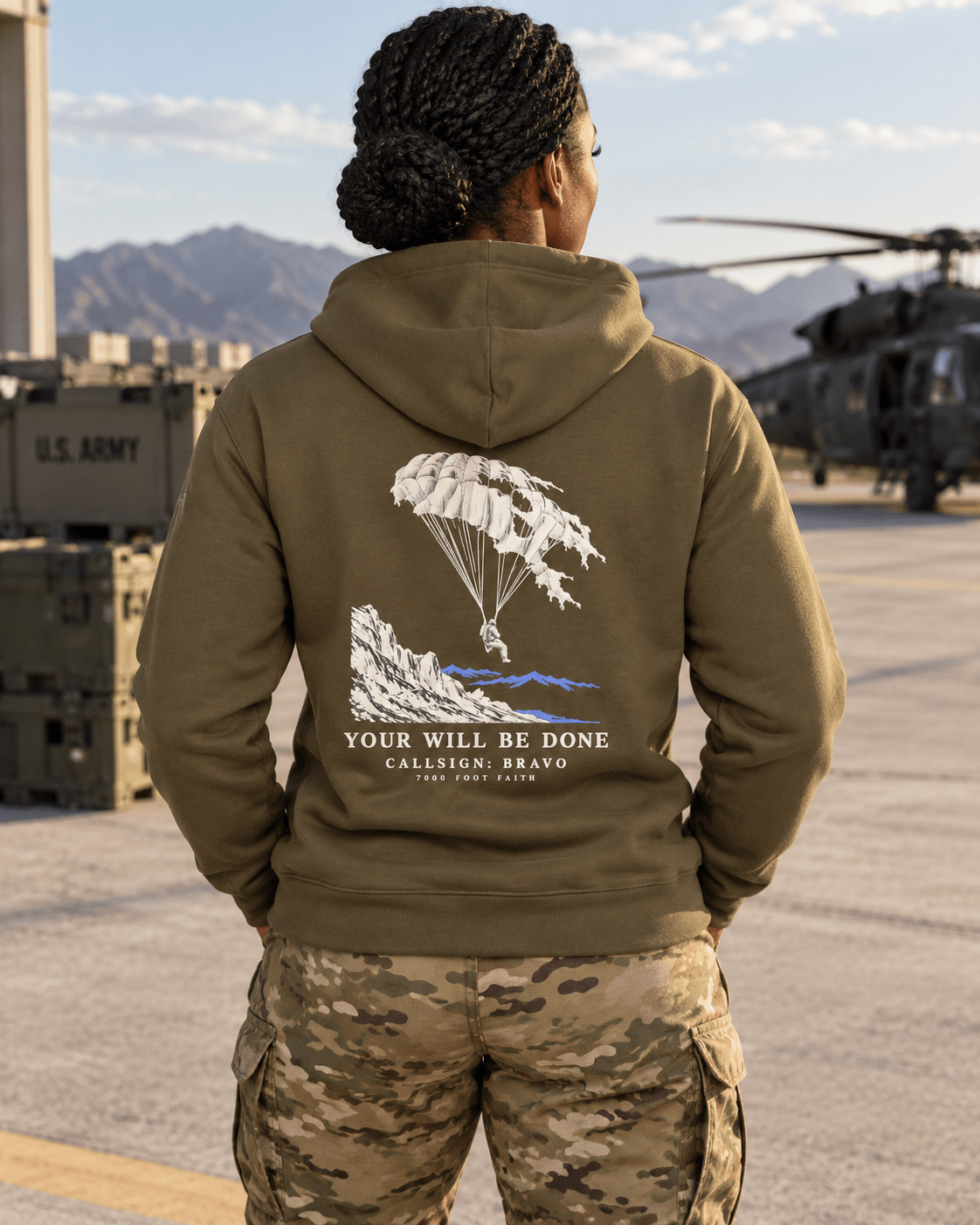 Woman wearing a Military Green Your Will Be Done: Descent CALLSIGN: BRAVO hoodie beside a helicopter on an airfield — back view