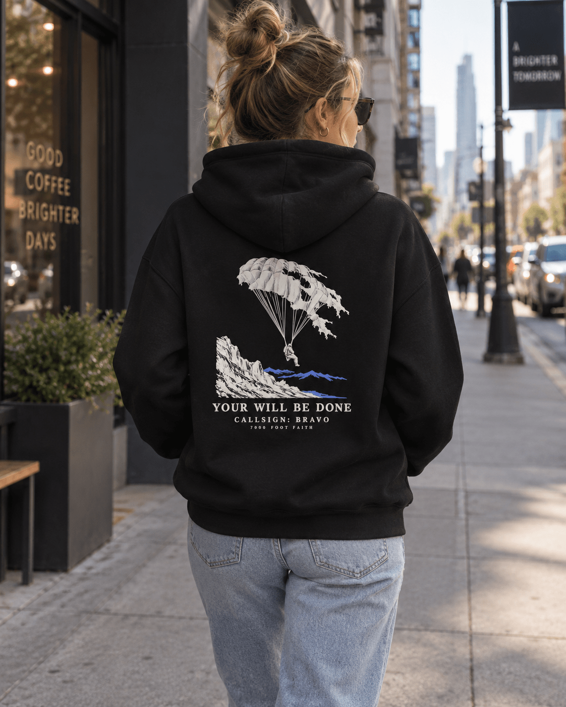 Woman wearing a Black Your Will Be Done: Descent CALLSIGN: BRAVO hoodie outside a city cafe — back view