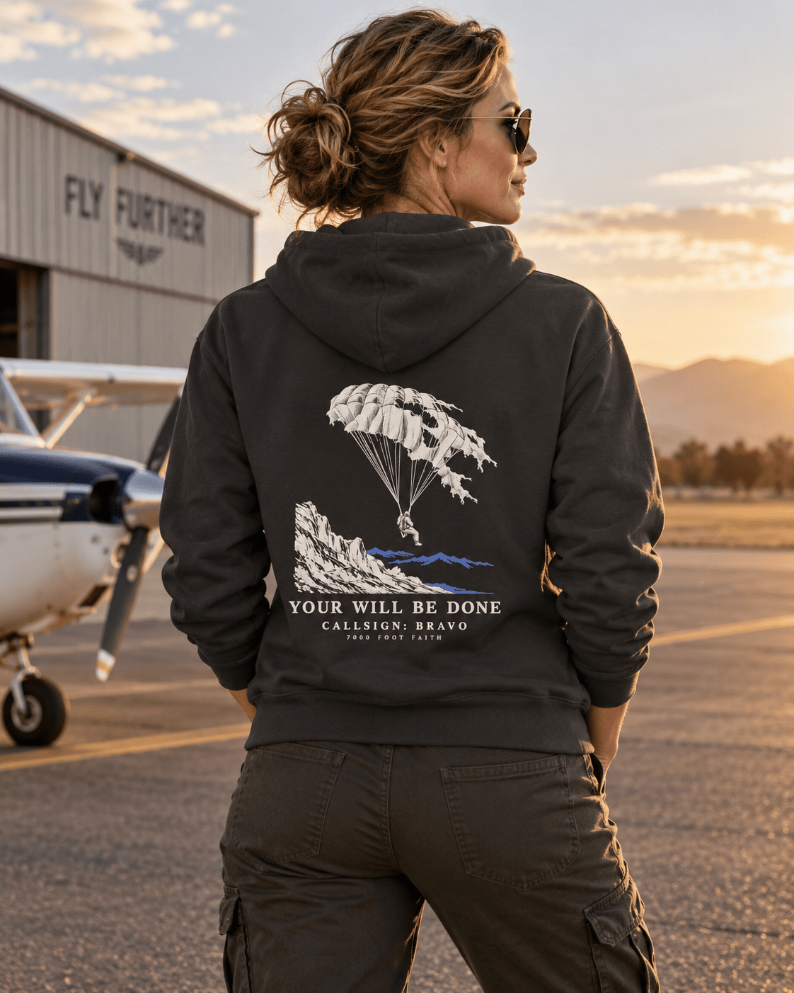 Woman wearing a Vintage Black Your Will Be Done: Descent CALLSIGN: BRAVO hoodie beside an aircraft hangar — back view