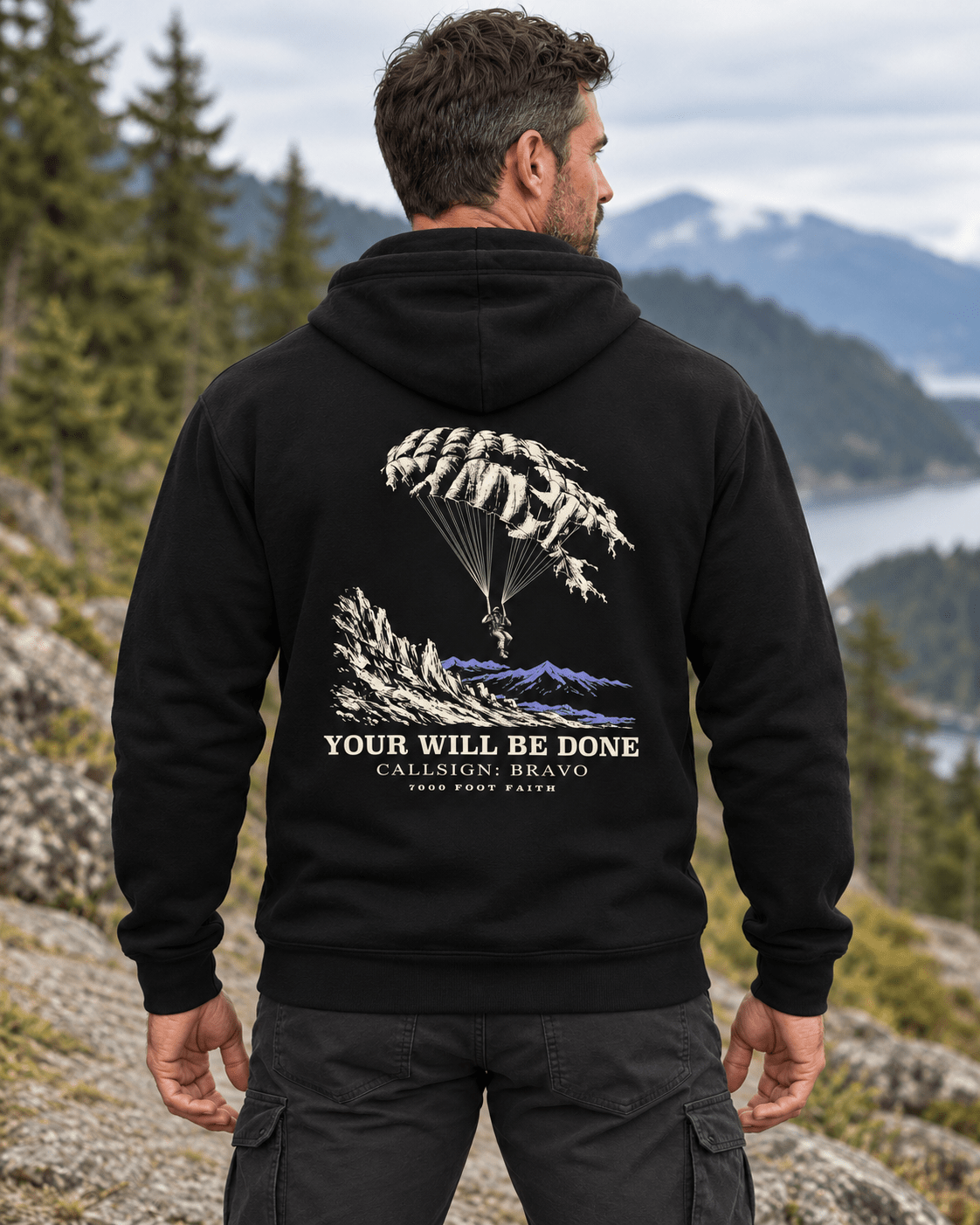 Man outdoors wearing a Black DESCENT faith hoodie, back view showing parachute and mountain artwork with Your Will Be Done lettering