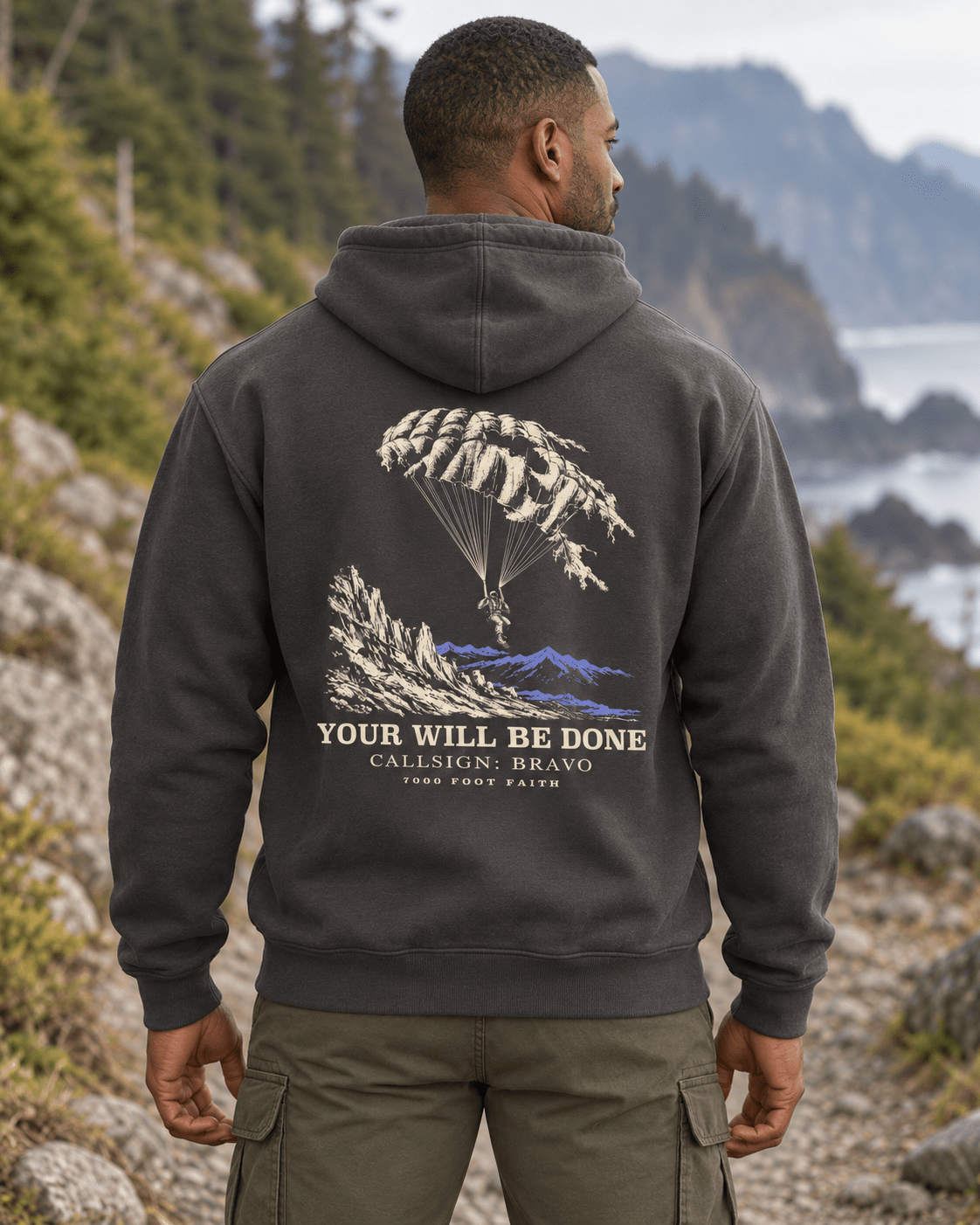 Man outdoors wearing a Vintage Black DESCENT faith hoodie, back view showing parachute and mountain artwork with Your Will Be Done lettering