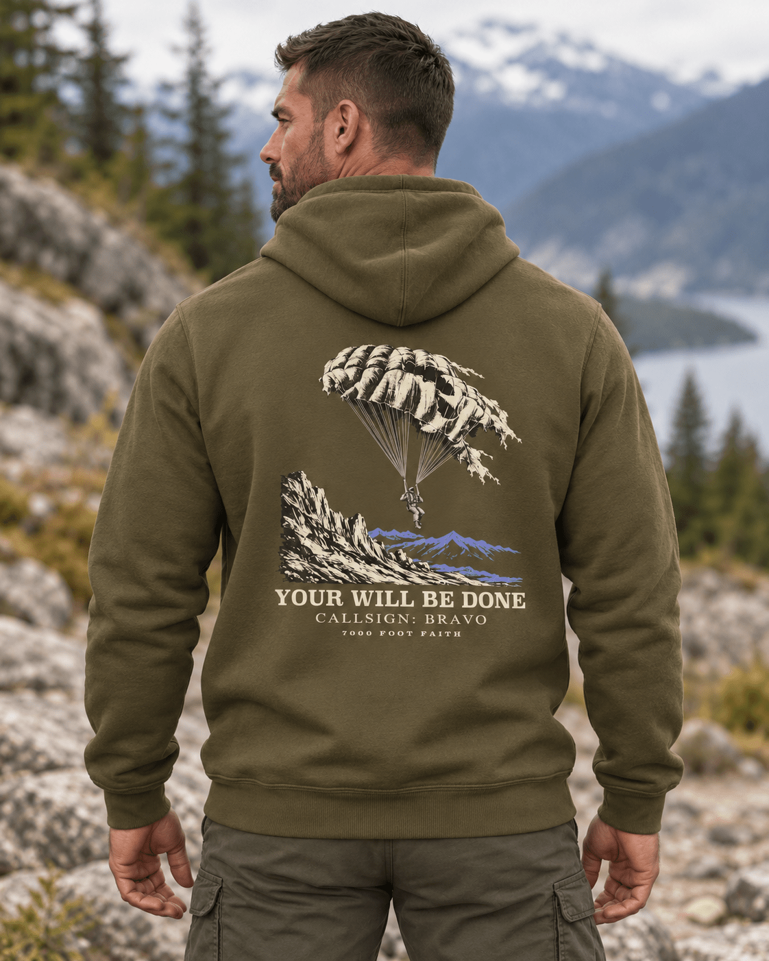 Man outdoors wearing a Military Green DESCENT faith hoodie, back view showing parachute and mountain artwork with Your Will Be Done lettering