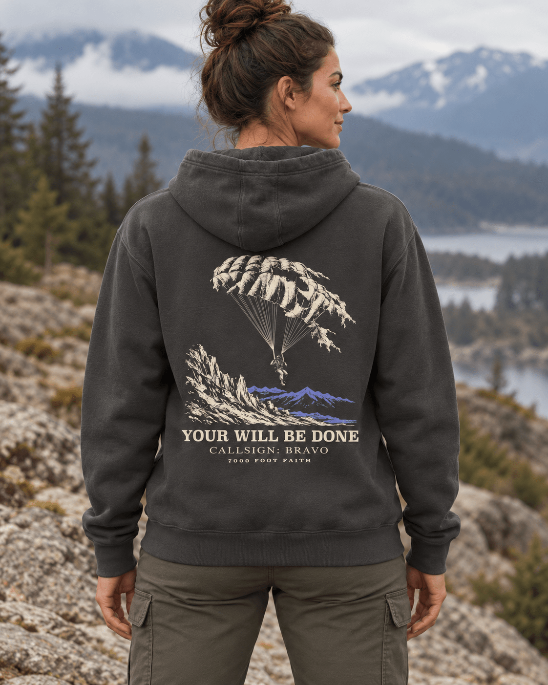 Woman outdoors wearing a Vintage Black DESCENT faith hoodie, back view showing parachute and mountain artwork with Your Will Be Done lettering